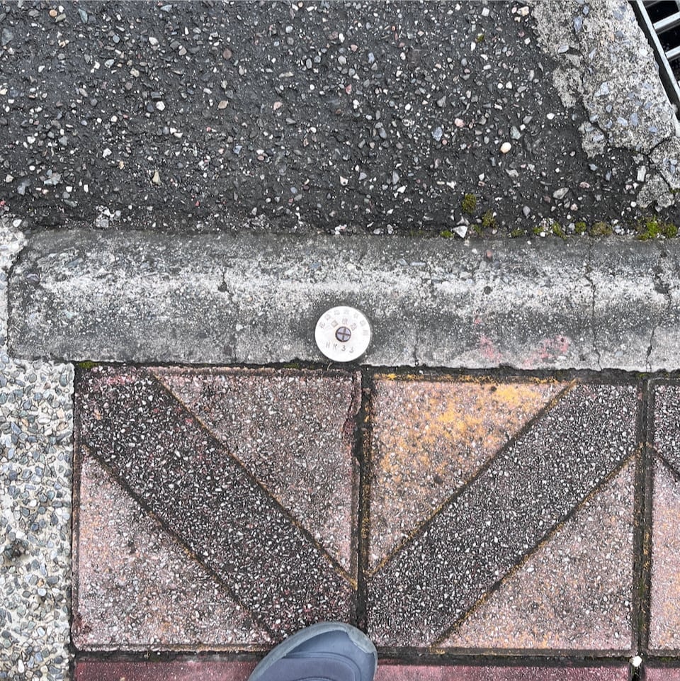 A photograph, looking directly down from a standing human's perspective. You can see a tiny bit of my shoe, and in the pavement is a circular metal plate about 5cm across. This one is marked `HK33`.